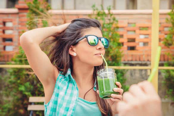 Beautiful young woman with sunglasses drinking green vegetable smoothie with straw in a summer day outdoors