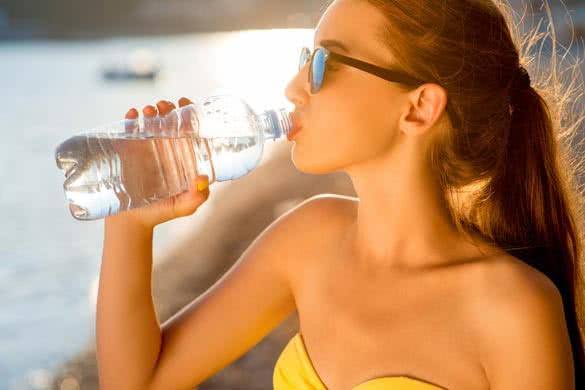 Young woman drinking sparkling water from transparent bottle on the beach on sunset