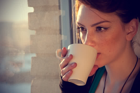 Beautiful young woman in casual wear dreaming with cup of hot tea over window