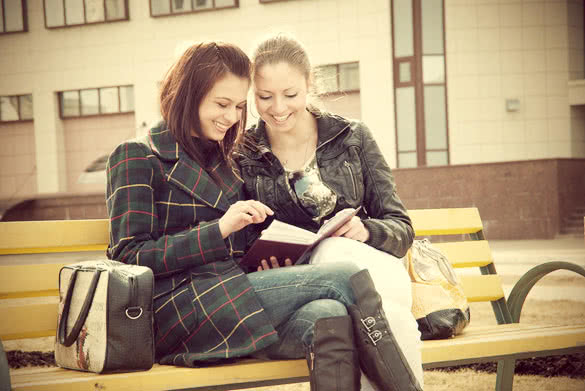 Two happy girls watch photos in album sitting on a bench outdoors