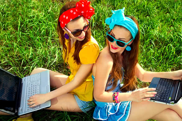 Two young stylish woman using laptops back to back on a green lawn