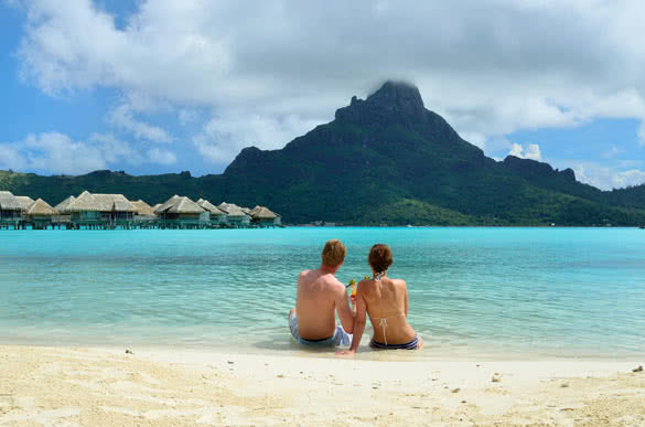 A honeymoon couple drinking a cocktail on the beach of a luxury vacation resort in the lagoon with a view on the tropical island of Bora Bora
