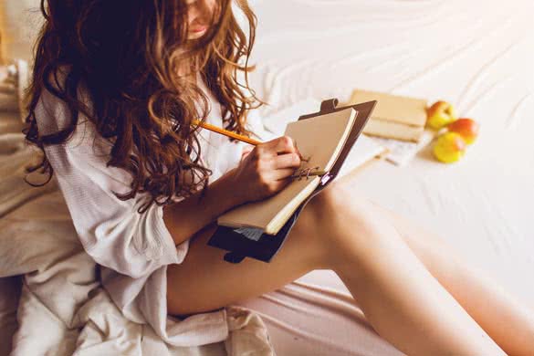 Close up lifestyle soft image of pretty young woman sitting on her cozy bed and making notes to her diary