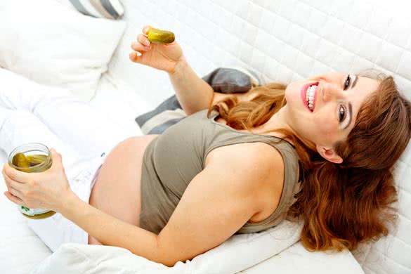 Smiling pregnant woman relaxing on sofa and holding jar of pickles