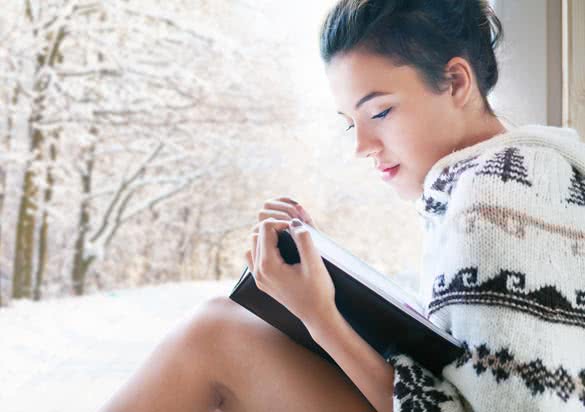 Beautiful young brunette woman reading book wearing knitted poncho sitting home by the window
