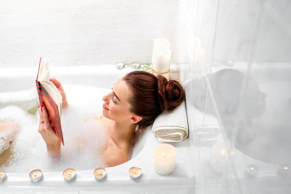 Young woman reading book while lying in the bath with foam and candles