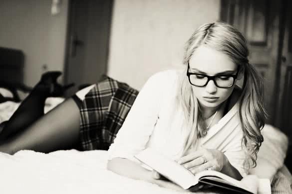 Sexy clever blonde woman in glasses reading book on bed