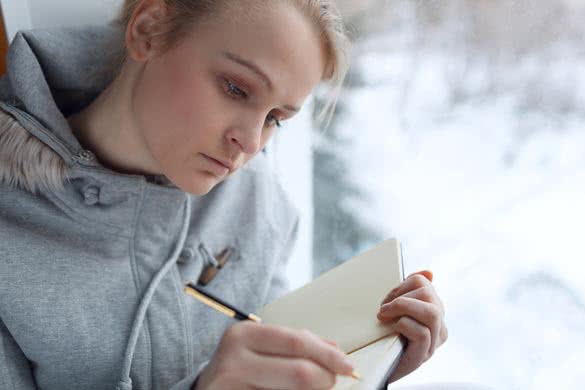 Young girl writing in her journal while sitting at a large window