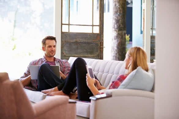 Couple At Home On Sofa In Lounge Using Digital Devices