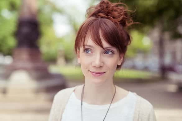 Pensive young redhead woman standing outdoors in an urban park looking to the side with a quiet smile and thoughtful expression