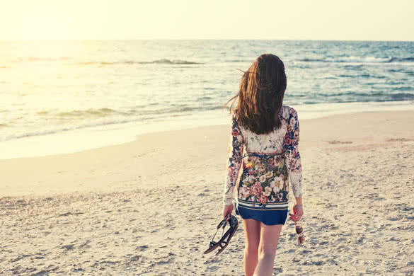 young and beautiful woman in colorful dress walking on the beach near the ocean and looking far away at the sunset