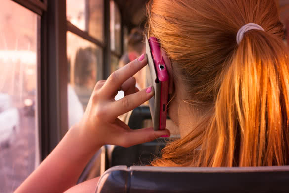 Attractive teenage girl with red orange hair talking over her pink smartphone in public transport looking through window in a sunsetting light
