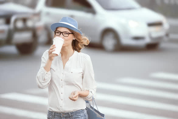 Happy young fashionable student drinking hot take away coffee and crossing a road against urban background