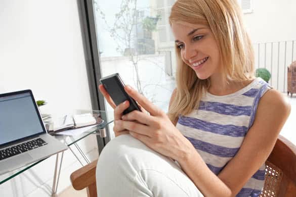 Portrait of a young beautiful woman sitting smiling in her home office space using a smartphone mobile phone