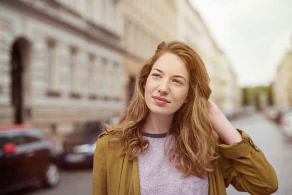 Attractive young woman standing in an urban street thinking deeply staring off to the side with a faraway expression and her hand to her hair