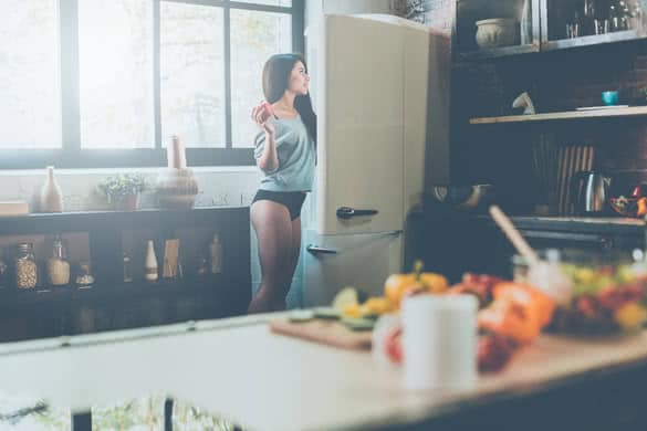 Beautiful young African woman looking into the fridge and holding red pepper while standing in kitchen at home