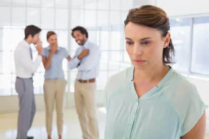 Colleauges gossiping with sad young businesswoman in foreground at a bright office