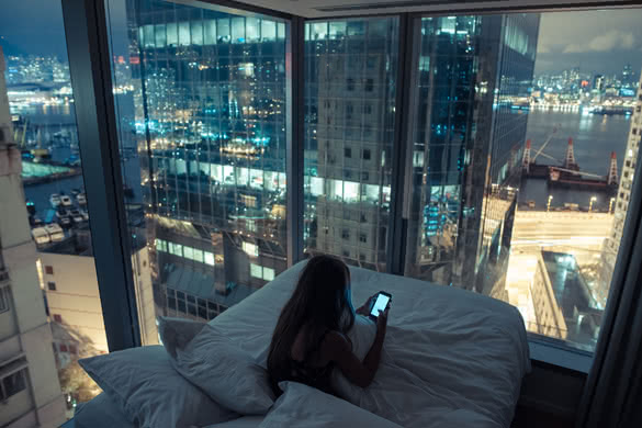 Young girl or teenager with long hair girl checking her mobile phone or chatting with someone at night in her bed with breathtaking view over night city