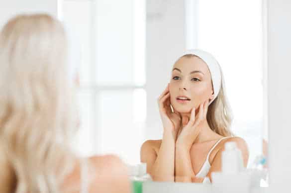 smiling young woman in hairband touching her face and looking to mirror at home bathroom