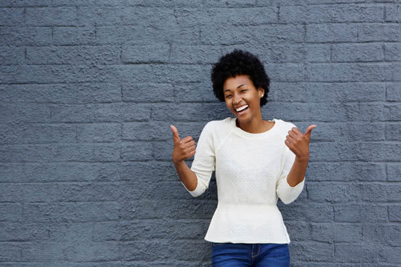 Portrait of smiling young african american woman gesturing thumbs up sign with both her hands against gray wall