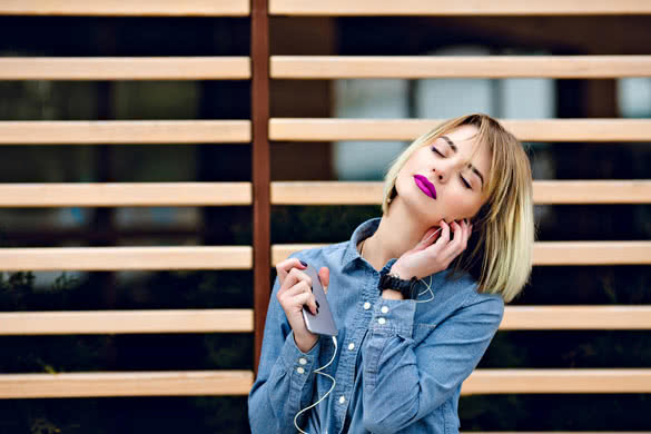 Stylish and bright girl with blonde hair and purple lipstick listening to music on the background of wooden wall