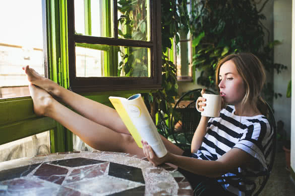 Stylish young woman in striped shirt sitting on beautiful european balcony drinking coffee and reading a book