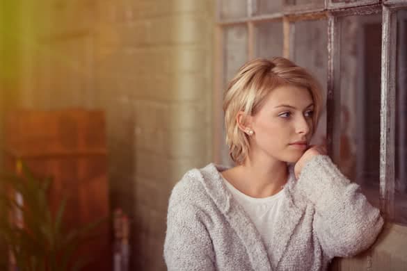 Young woman sitting daydreaming or waiting for someone staring out of a window in the living room with a serious expression