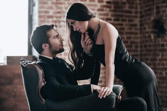 Beautiful young woman in cocktail dress leaning to her boyfriend sitting in chair while looking at each other in loft interior