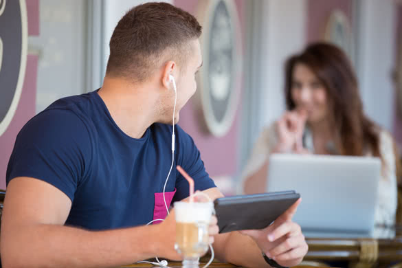 Handsome guy in casual clothes with tablet and earphones looking at smiling at him beautiful young woman sitting with a drink at the next table in street cafe