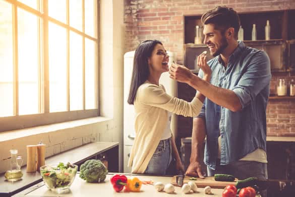 Beautiful young couple is feeding each other and smiling while cooking in kitchen at home