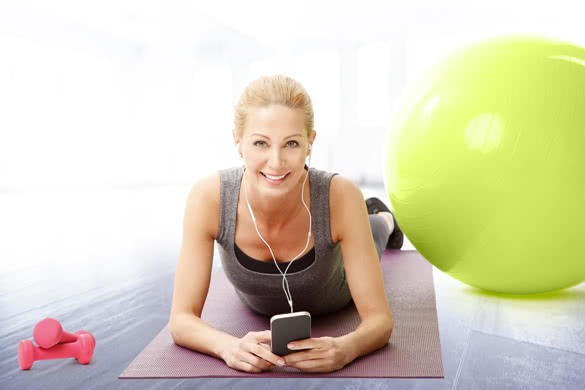 Portrait of smiling middle age woman lying on yoga mat after fitness workout next to exercise ball and listening music