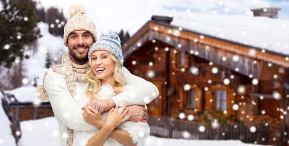 smiling man and woman in hats and scarf hugging over wooden country house and snow background