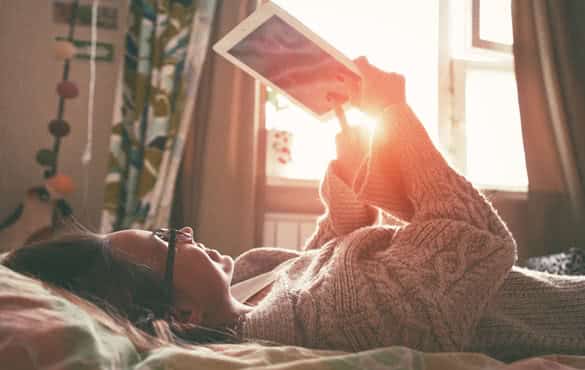 woman lying in bed with digital tablet touching with finger in morning sunlight